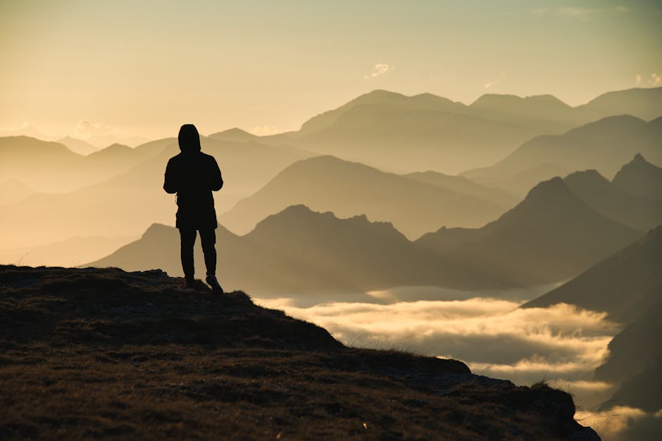 Silhouette of a person standing on a cliff with a stunning mountain view at dawn, surrounded by clouds.